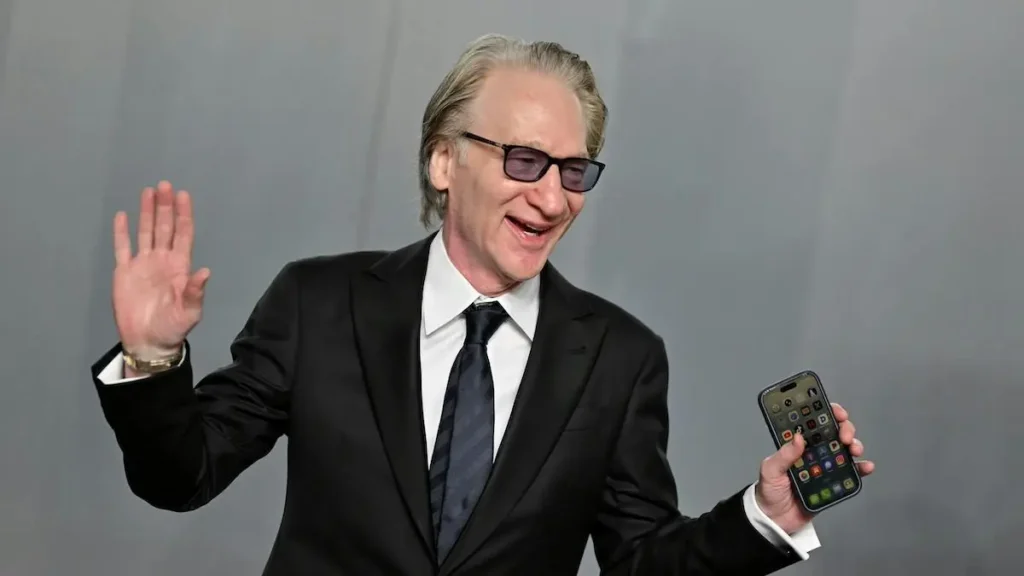 A man in a suit is waving and holding a smartphone, possibly at a public event or press conference.