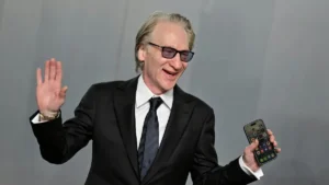 A man in a suit is waving and holding a smartphone, possibly at a public event or press conference.