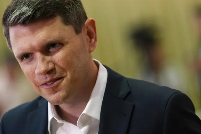 The image shows a man in a suit, appearing to be in a professional or formal setting.