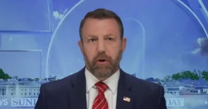The image shows a man in a suit and tie, speaking on a news program with a backdrop featuring the Washington Monument and the White House.