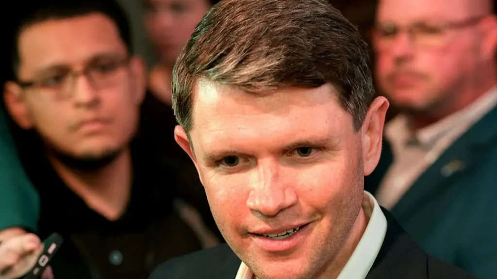 The image shows a man in a suit speaking at a press conference, with other individuals in the background.