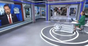 The image shows a news studio with a host seated at a desk, and a large screen displaying a segment titled "Face the Nation."