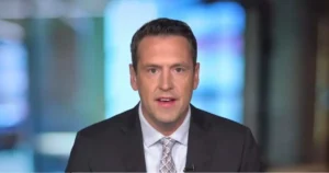 The image shows a news anchor in a professional setting, wearing a suit and tie, with a blurred background suggesting a studio environment.