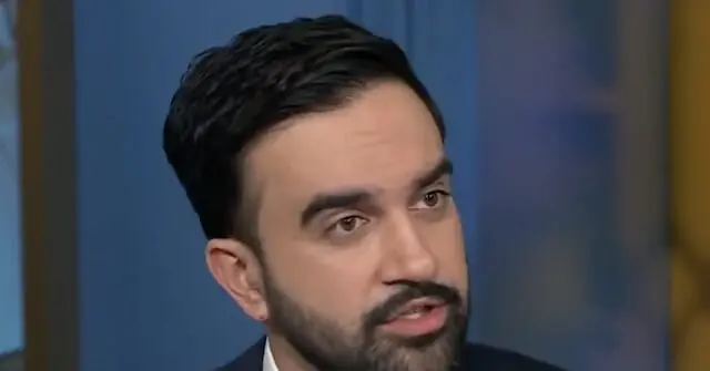 The image shows a man with a beard and mustache, wearing a suit, in what appears to be a news studio setting.