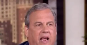The image shows a man speaking, with a blurred background that appears to be an indoor setting with architectural details.