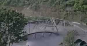 The image shows a damaged structure, possibly a bridge, with visible structural failure and debris scattered around, indicating a significant incident.