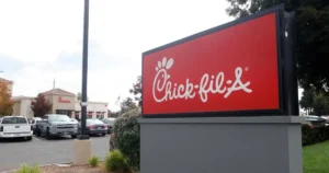 The image shows a Chick-fil-A sign in front of a restaurant with a parking lot full of cars.