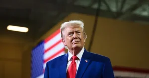 The image shows a person in a blue suit and red tie standing in front of an American flag.