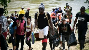 A group of people, some carrying bags, are walking along a riverbank, possibly after being rescued or relocating.