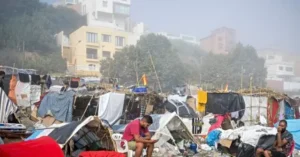 The image depicts a makeshift settlement with temporary shelters and laundry hanging, set against a backdrop of urban buildings and trees.