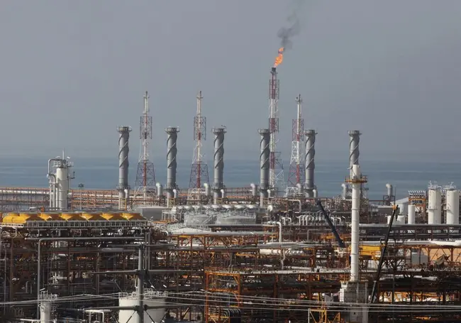 The image shows an industrial facility with tall towers and a flare stack emitting a flame, likely part of an oil or gas processing plant.