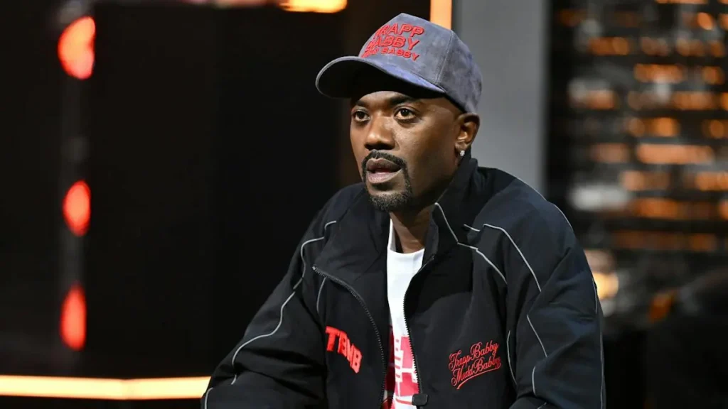The image shows a person wearing a black jacket with red text and a matching cap, sitting in a studio setting with blurred lights in the background.