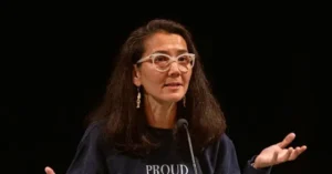 The image shows a person speaking at a podium, wearing a dark shirt with the word "PROUD" visible, and gesturing with their hands.