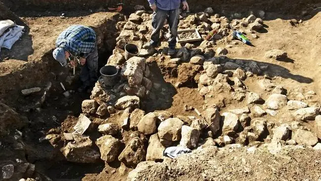 The photo depicts archaeologists working at an excavation site, carefully uncovering and examining ancient artifacts and structures.