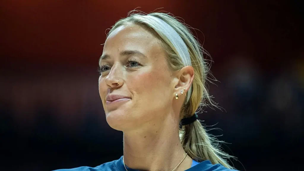The image shows a person with blonde hair tied back, wearing a headband and a blue top, smiling and looking to the side.