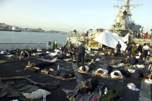 A group of sailors rests on blankets on the deck of a naval ship, with a large ship in the background and a cityscape visible in the distance.