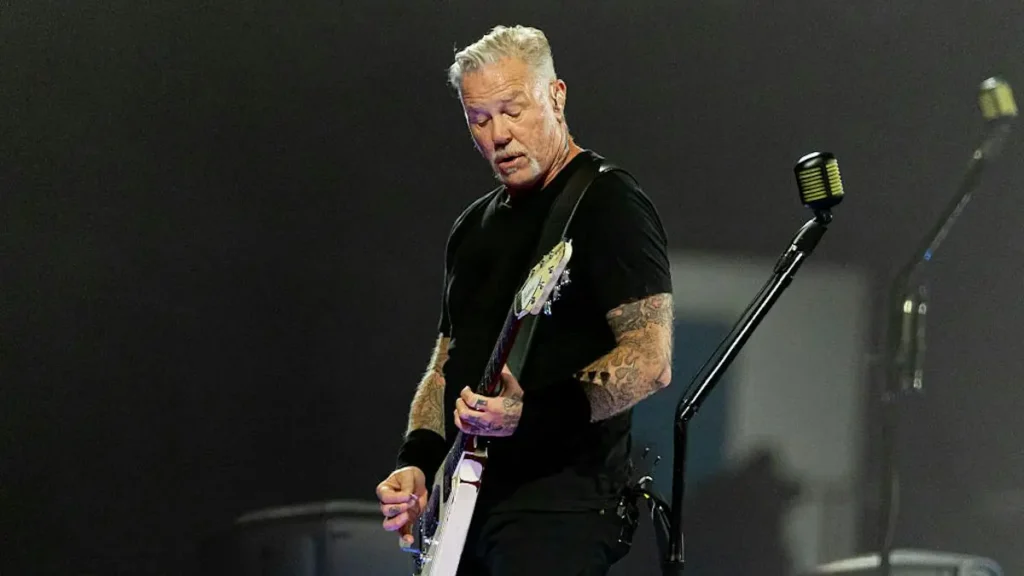 The image shows a musician performing on stage, holding a guitar and wearing a black shirt with visible tattoos on the arms.