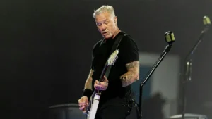 The image shows a musician performing on stage, holding a guitar and wearing a black shirt with visible tattoos on the arms.