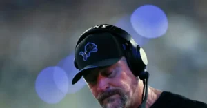 The image shows a person wearing a black cap with a logo and a headset, likely a coach or team official, in a sports setting.