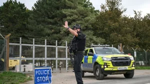 A police officer directs traffic near a "Police Stop" sign and a police vehicle, with a fenced area and trees in the background.