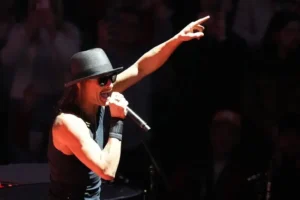 A performer in a black hat and sunglasses is passionately singing and pointing towards the audience during a live performance.
