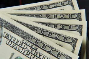 The image shows a close-up of several hundred-dollar bills fanned out, highlighting the intricate security features and denominations.