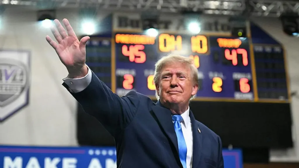 The image shows a person in a suit waving, with a scoreboard in the background displaying a basketball game score.
