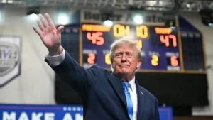 The image shows a person in a suit waving, with a scoreboard in the background displaying a basketball game score.