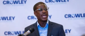 A man in a suit and glasses is speaking into a microphone at a press event with a backdrop featuring the "Crowley" logo.