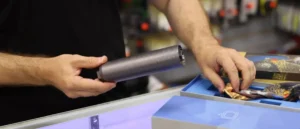 A person is using a cylindrical tool to interact with a box containing various items, possibly in a retail or demonstration setting.
