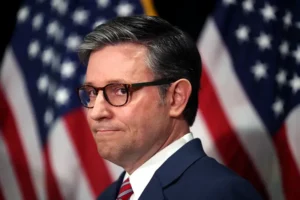 The image shows a man in a suit and tie, wearing glasses, standing in front of American flags.