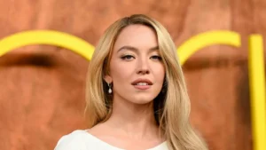The image shows a person with long blonde hair wearing a white top and diamond earrings, standing in front of a backdrop with yellow and brown tones.