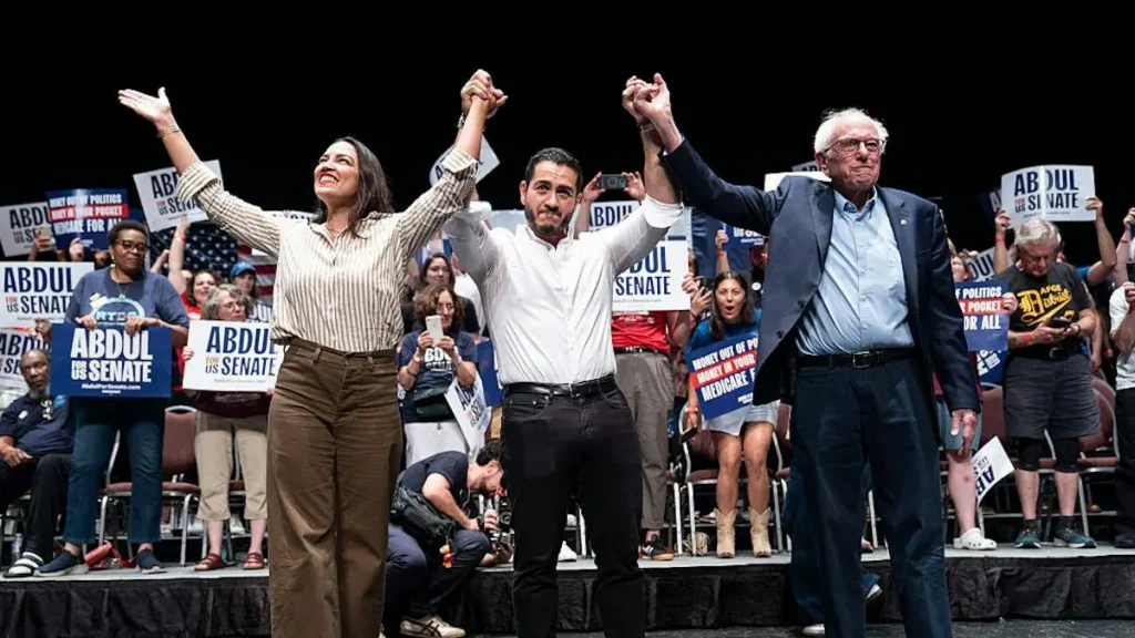 A political rally features three individuals on stage, raising their arms in unity, with supporters holding signs in the background.
