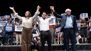 A political rally features three individuals on stage, raising their arms in unity, with supporters holding signs in the background.