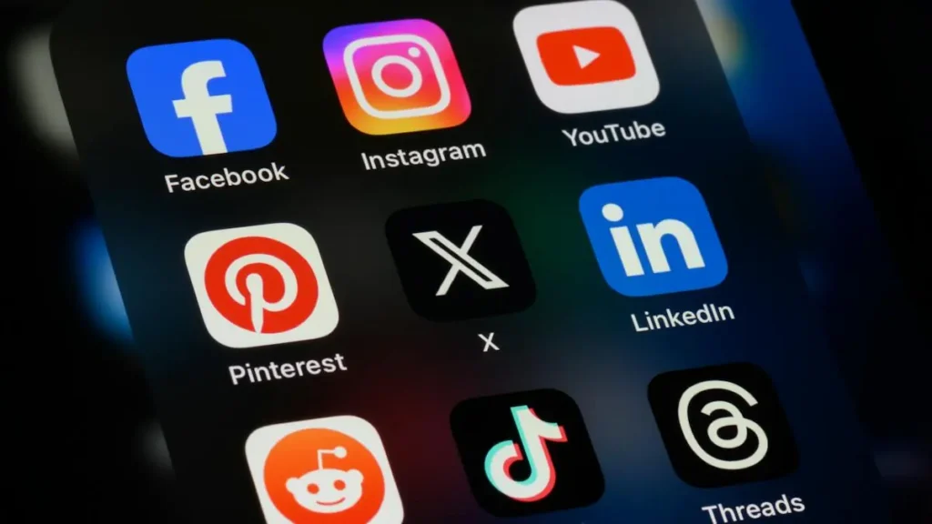 The image shows a smartphone screen displaying various social media app icons, including Facebook, Instagram, YouTube, Pinterest, X (formerly Twitter), LinkedIn, Reddit, TikTok, and Threads.