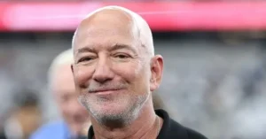 The image shows a man with a bald head and a light beard, smiling, with a blurred background that appears to be an outdoor setting.