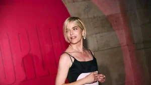 The image shows a person wearing a black sleeveless top and pearl jewelry, standing in front of a red and beige backdrop.