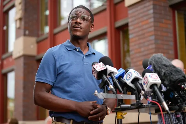 A man in a blue shirt stands at a press conference with microphones from various news outlets.