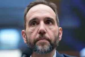 The image shows a man with a beard and short hair, wearing a suit, speaking into a microphone in what appears to be a formal setting.