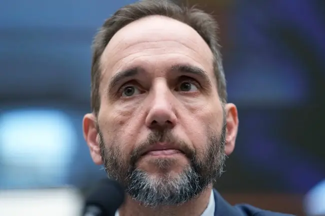 The image shows a man with a beard and short hair, wearing a suit, speaking into a microphone in what appears to be a formal setting.