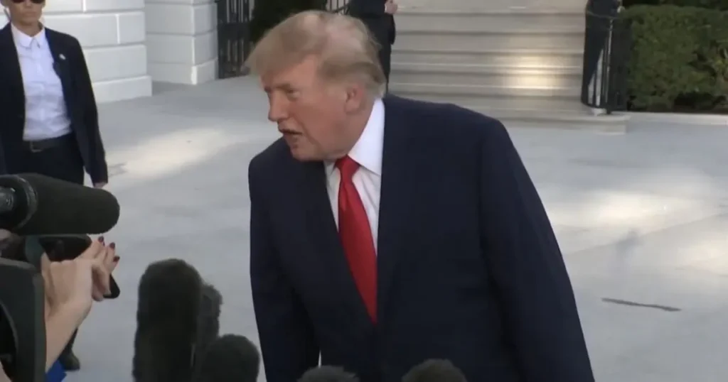 The image shows a man in a suit and red tie speaking to reporters in front of a building with steps.