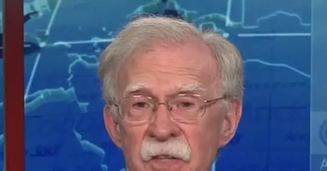 The image shows a man with gray hair and a mustache, wearing glasses, speaking in front of a blue background with a map.