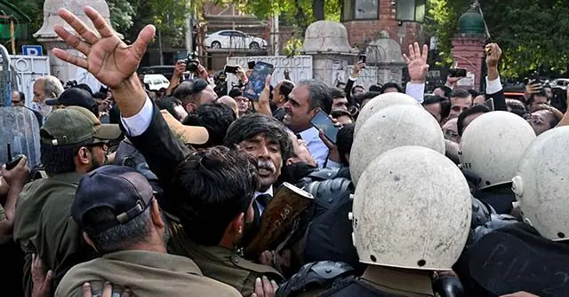 A man raises his hands in a crowd of police officers and protesters during a tense confrontation.