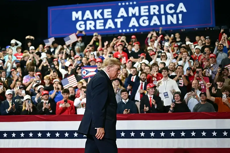 The image shows a political rally with a crowd enthusiastically cheering and waving flags, with a prominent sign reading "MAKE AMERICA GREAT AGAIN!" in the background.