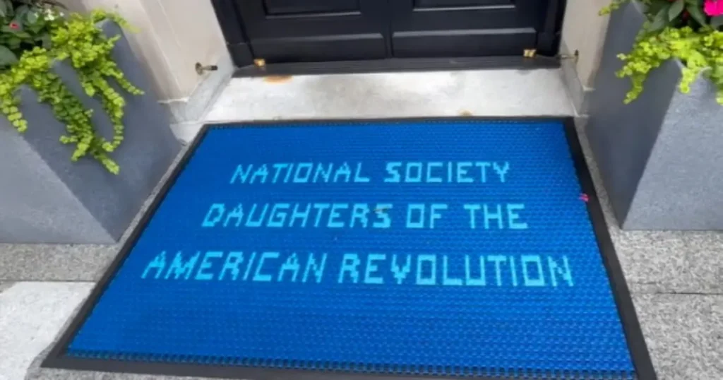 The image shows a blue doormat with the text "NATIONAL SOCIETY DAUGHTERS OF THE AMERICAN REVOLUTION" in front of a door.