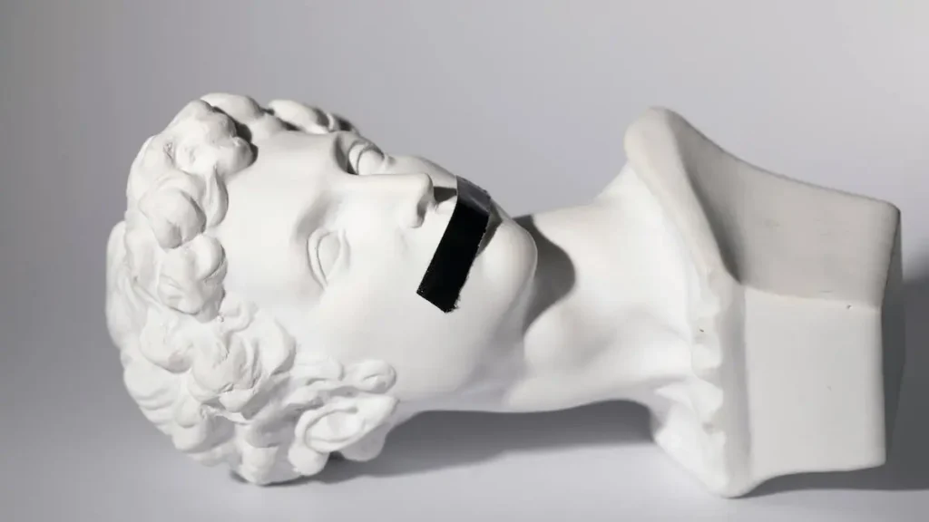 A classical sculpture of a head with a black tape covering its mouth is displayed against a plain background.