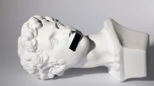 A classical sculpture of a head with a black tape covering its mouth is displayed against a plain background.