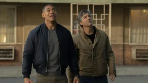 Two individuals are walking side by side, looking up, in an urban setting with a building in the background.