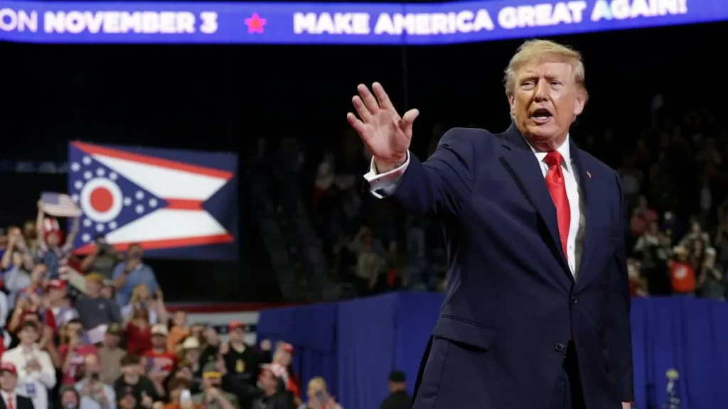 The image shows a political rally with a prominent figure in a suit and tie, addressing a crowd, with a "Make America Great Again" banner and an Ohio state flag in the background.