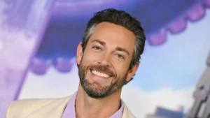 The image shows a man with a beard and short hair, smiling, wearing a light-colored blazer over a purple shirt, with a colorful background.